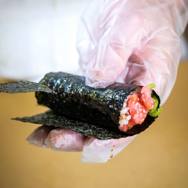 Gloved hands preparing a Sweetfish sushi roll with tuna, rice, and green lettuce.