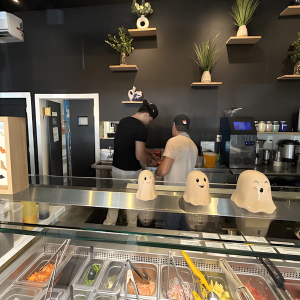 Two Sweetfish Poké Bowls & Sushi employees prepare food behind a counter with ingredients and ghost decorations.