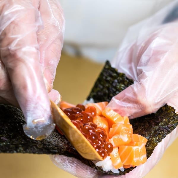 Gloved hands preparing a Sweetfish sushi roll with salmon, roe, and rice on nori.