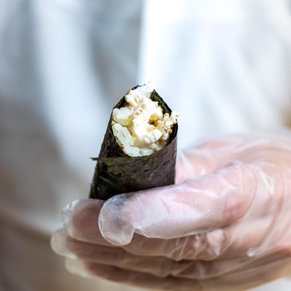 A gloved hand holds a Sweetfish Poké Bowls & Sushi hand roll filled with rice and creamy seafood.