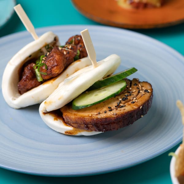 Two Sweetfish bao buns on a blue plate, one with pork belly and cucumber, the other with chicken.